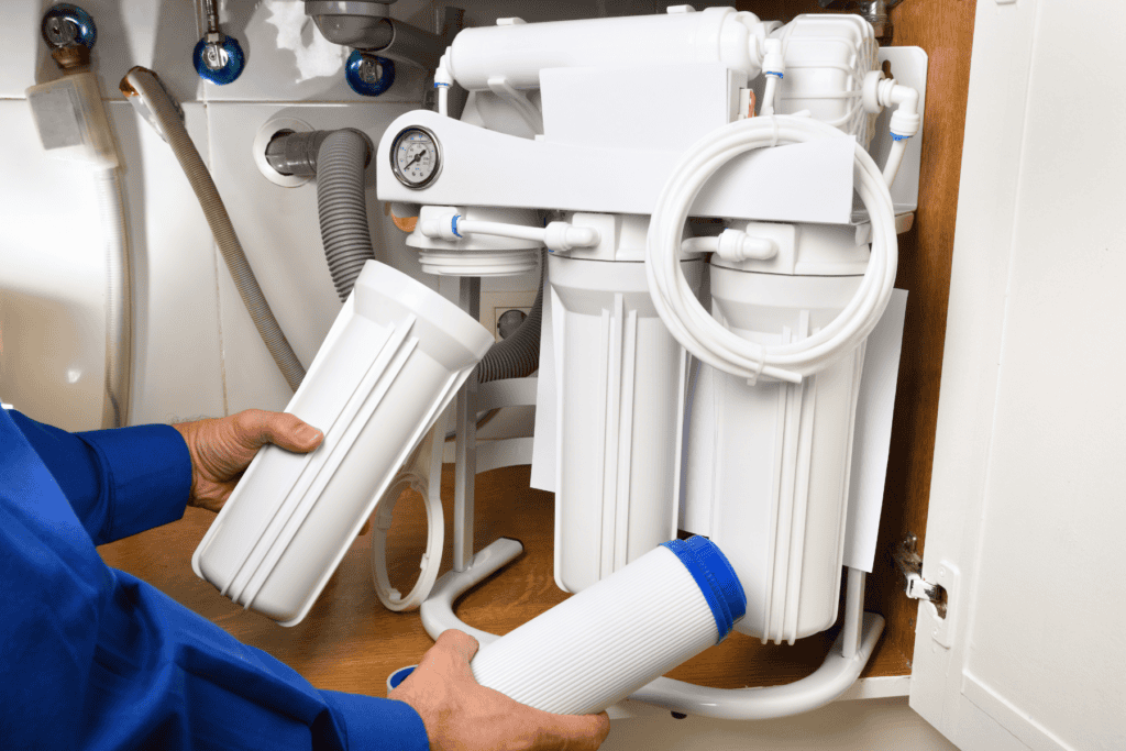Person replacing a filter in an under-sink reverse osmosis water filtration system with multiple canisters and a pressure gauge.