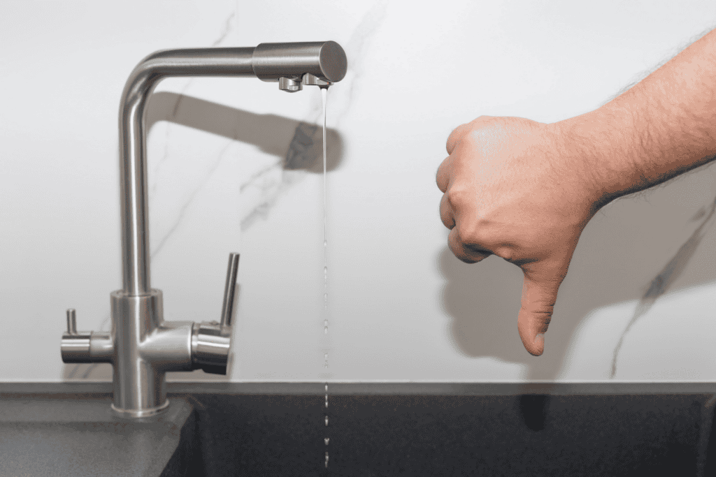 A hand showing a thumbs-down next to a kitchen faucet with a weak stream of water coming out.