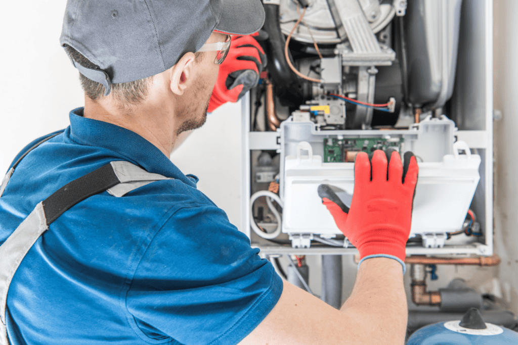 A technician wearing red gloves and a blue shirt repairs or inspects an open boiler or heating unit, exposing internal electronic components and wiring.